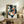 May include: Framed artwork displaying a decorative koi fish with coral and cream colored fins swimming among stylized ginkgo leaves against a dark navy dotted background, positioned on a rustic wooden sideboard next to a fern plant, books, and ceramic vessels in a neutral-toned living room setting.