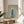 May include: Framed poster featuring a cream-colored poodle wearing a teal knit sweater and small boots, styled like a fashion magazine cover with "DOGUE" in gold letters at the top. The wooden frame sits on a round wooden side table next to a beige upholstered chair, with natural sunlight streaming through white curtains and a plant visible in the corner of this bright, modern living room setting.