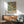 May include: William Morris exhibition poster featuring orange fruit and green botanical pattern hanging in wood-framed display above mid-century modern sideboard. Scene shows sage green upholstered armchair with wooden arms, round coffee table, and white ceramic table lamp. Warm natural lighting creates geometric shadows on white wall in contemporary living room setting.
