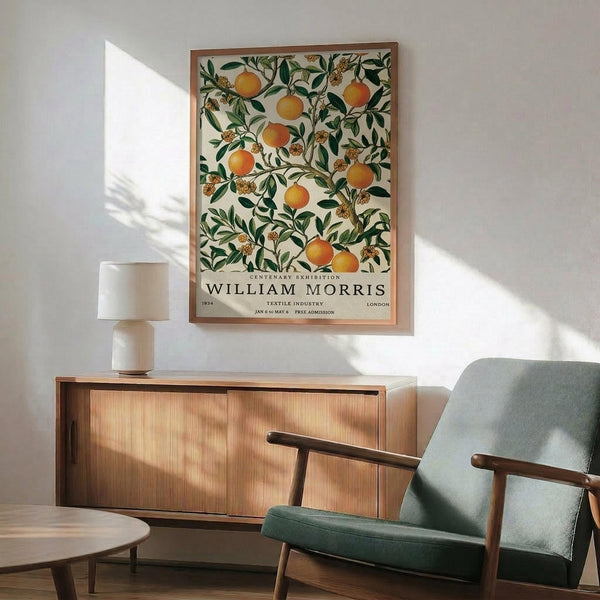 May include: William Morris exhibition poster featuring orange fruit and green botanical pattern hanging in wood-framed display above mid-century modern sideboard. Scene shows sage green upholstered armchair with wooden arms, round coffee table, and white ceramic table lamp. Warm natural lighting creates geometric shadows on white wall in contemporary living room setting.