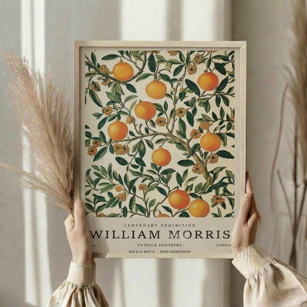 May include: Person holding framed botanical poster featuring orange citrus fruits and green foliage in William Morris decorative style against cream background, with dried pampas grass stems visible to left, neutral curtained interior setting