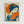 A Japanese-style art print displayed in a light wood frame on a white wall. The artwork features a geisha with dark blue hair adorned with orange flowers, holding a bright orange folding fan that partially covers her face. She wears a blue kimono with wave patterns. The composition uses a limited color palette of orange, blue, and cream with Japanese characters in the upper left corner. The print has a vintage poster aesthetic with visible texture and grain.