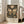May include: Framed vintage-style exhibition poster featuring large ornate butterfly with cream and olive green wings surrounded by detailed botanical flowers and foliage on dark background. White matted frame leans against white wall next to beige upholstered sofa in bright sunlit room with hardwood floors.