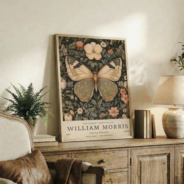 May include: Framed botanical poster featuring large butterfly with circular wing patterns surrounded by flowers and foliage in muted greens, peaches, and creams. Exhibition-style text at bottom. Displayed on rustic wooden sideboard with books, ceramic vase, and fern plant. Cream colored armchair with leather pillow visible in cozy living room setting with natural lighting.