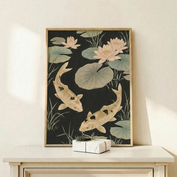 May include: Framed Japanese-style artwork showing two golden koi fish swimming among pink lotus flowers and green lily pads on a dark background, displayed on a light wood console table next to a small white wrapped gift with greenery accent against a cream wall.