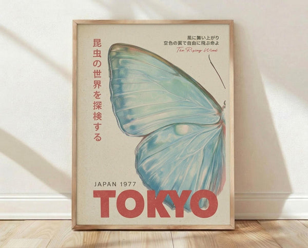 May include: Framed vintage-style poster with light blue butterfly wing design against cream background, featuring red Japanese text vertically aligned on left side, "The Rising Wind" in script, and bold red "TOKYO" text at bottom with "JAPAN 1977". Natural wood frame displayed on light wooden floor against white wall with soft shadows.