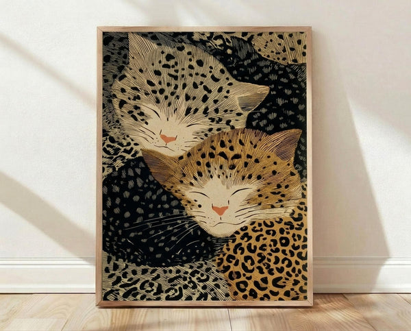 May include: Framed art print featuring two stylized cats with human-like faces in a Japanese woodblock print style. One cat has spotted leopard-like markings in golden brown, the other has black and white coloring. Both cats have peaceful, smiling expressions with closed eyes and pink noses. The artwork shows intricate line work and traditional Japanese artistic techniques against a dark background. The print is displayed in a light wooden frame, leaning against a white wall on light wooden flo