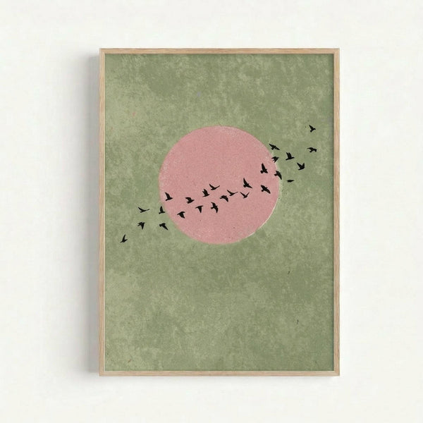 Minimalist art print featuring a large pink circle resembling a sun against a mottled sage green background, with a flock of small black bird silhouettes flying diagonally across and around the circle. The artwork is displayed in a light wooden frame hanging on a white wall, creating a modern, serene aesthetic with natural earth tones.