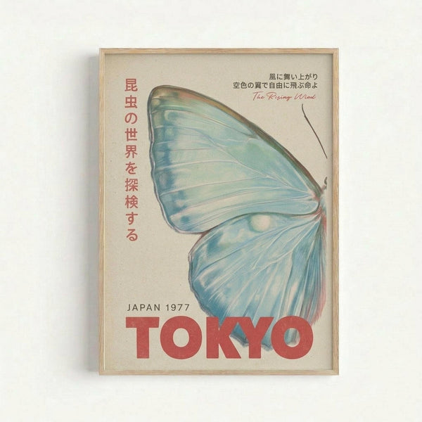 Framed vintage-style travel poster featuring a large blue butterfly wing with delicate shading and veining. Japanese text in red characters runs vertically down the left side, with additional Japanese text and "The Rising Wind" in script at the top right. Bold red "TOKYO" text spans the bottom with "JAPAN 1977" above it. The poster has a cream background and is displayed in a light wooden frame against a white wall.