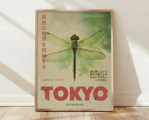 May include: Framed vintage-style poster with green dragonfly illustration on cream background with Japanese text down left side, red TOKYO text at bottom, and Japan 1977 date. Wood frame sits on light wooden floor against white wall with soft shadows.