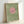 Minimalist poster print featuring a large pink circle on a textured sage green background with black bird silhouettes flying in formation across and around the circle. The artwork is displayed in a light wood frame leaning against a white wall, showing the simple geometric design and nature-inspired elements in muted, earthy tones.