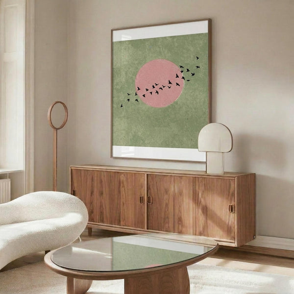 May include: Minimalist poster with green textured background featuring pink circular sun and black silhouettes of birds in flight, displayed in thin brown frame above wooden sideboard with brass hardware. White curved chair and glass-top coffee table visible in modern living room with cream walls and natural lighting.
