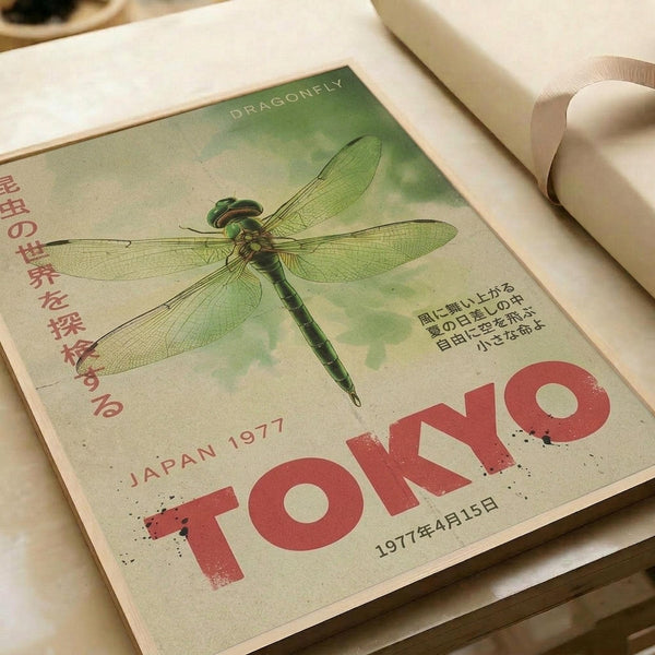 May include: Vintage-style poster print featuring a detailed green dragonfly illustration on cream background with red "TOKYO" text and "JAPAN 1977" marking. Japanese characters appear on left side. Print shows weathered, aged texture and sits on white fabric surface next to rolled papers, creating an artistic flat lay scene.