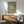 May include: Framed poster with green textured background, large pink circle resembling sun or moon, and flock of small black birds flying across. Wood-framed artwork displayed above mid-century modern wooden credenza with table lamp. Sage green upholstered chair with wooden arms visible in foreground. Warm natural lighting creates shadows on white wall in minimalist living room setting.