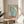 May include: A framed vintage-style poster featuring a large blue butterfly wing and Japanese text, displayed on a round wooden side table next to a beige upholstered chair. The poster has "JAPAN 1977" and "TOKYO" in red text with "The Rising Wind" in smaller script. Natural sunlight casts shadows across the neutral-toned room setting with a plant visible in the background.