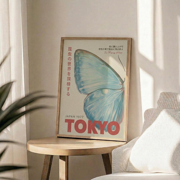 May include: A framed vintage-style poster featuring a large blue butterfly wing and Japanese text, displayed on a round wooden side table next to a beige upholstered chair. The poster has "JAPAN 1977" and "TOKYO" in red text with "The Rising Wind" in smaller script. Natural sunlight casts shadows across the neutral-toned room setting with a plant visible in the background.