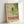 May include: Framed vintage-style poster with green dragonfly illustration on cream background, featuring Japanese text and "TOKYO" in red letters, "JAPAN 1977" and date "1977年4月15日", displayed in light wood frame leaning against white wall on light surface.