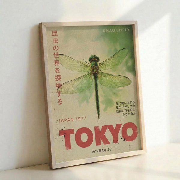 May include: Framed vintage-style poster with green dragonfly illustration on cream background, featuring Japanese text and "TOKYO" in red letters, "JAPAN 1977" and date "1977年4月15日", displayed in light wood frame leaning against white wall on light surface.