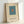 Minimalist art print featuring a teal blue rectangle with small orange square in center on cream background, displayed in light wood frame leaning against white wall with soft natural lighting
