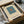May include: Minimalist abstract art print in black frame featuring teal square with small orange square center on cream background. Packaged in brown cardboard box with white tissue paper padding, displayed alongside wrapped cylindrical items on white fabric surface with glass vessel partially visible.