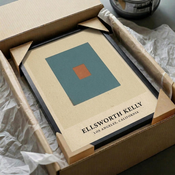 May include: Minimalist abstract art print in black frame featuring teal square with small orange square center on cream background. Packaged in brown cardboard box with white tissue paper padding, displayed alongside wrapped cylindrical items on white fabric surface with glass vessel partially visible.