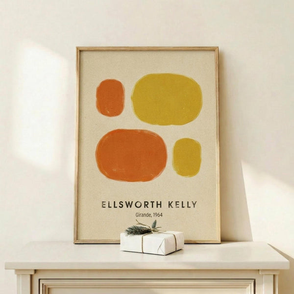 May include: Framed abstract art print with four organic oval shapes in orange and yellow tones on cream background, displayed on light wood console table. Small wrapped gift box with evergreen sprig sits in front of frame. Minimalist room setting with white walls and natural lighting.