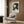 May include: Framed vintage Japanese exhibition poster with green and cream geometric umbrella design hanging on wood-paneled wall above beige upholstered armchair with patterned pillow. Modern floor lamp with brass finish and marble base stands nearby next to dark round side table with open book. Neutral tones throughout minimalist interior setting.
