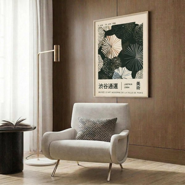 May include: Framed vintage Japanese exhibition poster with green and cream geometric umbrella design hanging on wood-paneled wall above beige upholstered armchair with patterned pillow. Modern floor lamp with brass finish and marble base stands nearby next to dark round side table with open book. Neutral tones throughout minimalist interior setting.
