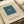 Minimalist art print featuring a teal blue square with a small orange square centered inside, displayed on cream-colored paper with black text reading "ELLSWORTH KELLY LOS ANGELES, CALIFORNIA" below the geometric design. The print is shown flat alongside rolled paper tubes on a neutral surface.