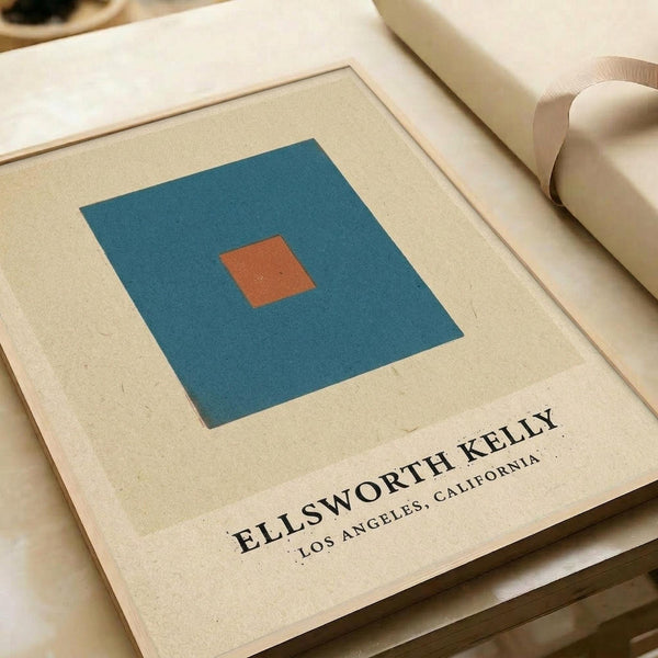 Minimalist art print featuring a teal blue square with a small orange square centered inside, displayed on cream-colored paper with black text reading "ELLSWORTH KELLY LOS ANGELES, CALIFORNIA" below the geometric design. The print is shown flat alongside rolled paper tubes on a neutral surface.