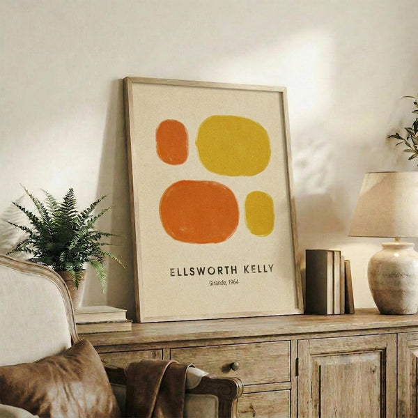 May include: Framed minimalist abstract art print featuring four organic oval shapes in orange and yellow tones on cream background, displayed on rustic wooden sideboard alongside fern plant, ceramic vases, and leather chair in warm-toned living room setting with natural lighting.