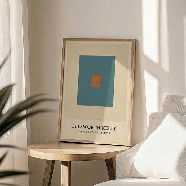 May include: Framed geometric art print with teal blue square containing small orange rectangle on cream background, displayed on round wooden side table next to beige upholstered chair. Natural sunlight streams across white walls, with green plant leaves visible in corner of minimalist living room setting.