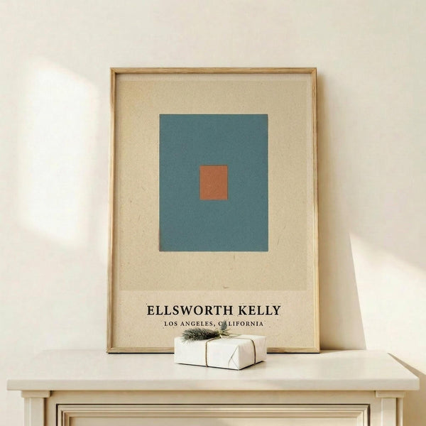 May include: Framed minimalist art print featuring a teal blue square with small orange square in center on cream background, displayed on light wood console table next to small wrapped gift with evergreen sprig, against white wall with natural lighting