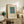 May include: Framed minimalist art print with teal blue rectangle and small orange square on cream background, displayed on rustic wooden sideboard alongside fern plant, ceramic vase, and stacked books in warm-toned living room setting with beige upholstered furniture.