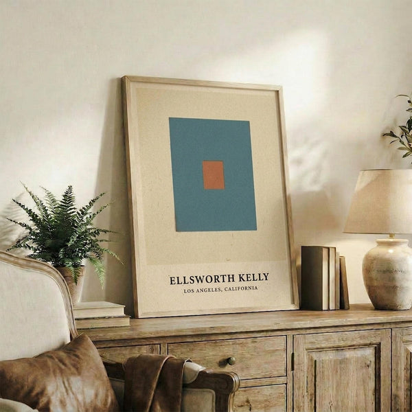 May include: Framed minimalist art print with teal blue rectangle and small orange square on cream background, displayed on rustic wooden sideboard alongside fern plant, ceramic vase, and stacked books in warm-toned living room setting with beige upholstered furniture.
