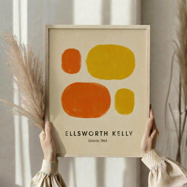 May include: Person holding framed abstract art print featuring four organic oval shapes in orange and yellow tones on cream background, displayed in light wooden frame. Dried pampas grass visible to left side. Neutral beige curtains and soft natural lighting create minimalist home decor setting.