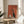 May include: Framed retro-style poster featuring a sleek black cat with white chest sitting beside a white coffee pot and cup on an orange-red background, displayed on a round wooden side table next to a beige upholstered chair, with plant leaves visible and natural sunlight casting shadows across the scene.