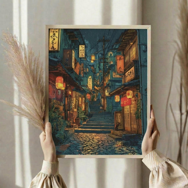 May include: Person in beige sweater holding framed artwork showing Japanese street scene with stone steps, traditional wooden buildings, glowing red lanterns, shop signs with Japanese characters, and rainy evening atmosphere in blue and warm golden tones. Dried pampas grass visible beside figure against white curtained background.