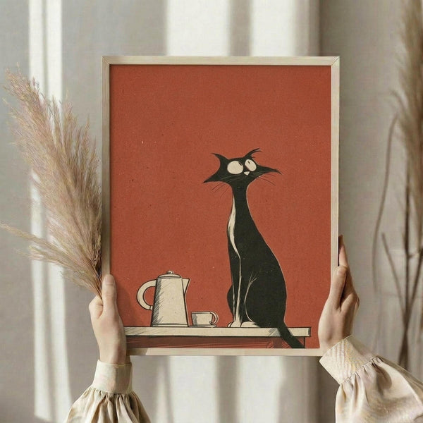 May include: Person holding framed vintage-style poster with orange-red background featuring stylized black cat with large white eyes sitting beside coffee pot and cup on wooden shelf. Light wood frame, dried pampas grass visible, neutral beige clothing and curtains in soft natural lighting.