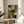 May include: Abstract geometric artwork in earth tones displayed in thin wooden frame on round natural wood side table. Composition features overlapping curved and angular shapes in various shades of green, olive, cream and dark forest green with textured, weathered appearance. Scene shows bright natural lighting, plant shadows, and neutral beige upholstered chair in modern minimalist interior setting.