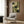 May include: Abstract geometric poster with green, cream, and dark color blocks and curved shapes, displayed in light wood frame on wood paneled wall above beige armchair with patterned pillow, brass floor lamp, and dark round side table with open book in modern neutral living room setting.