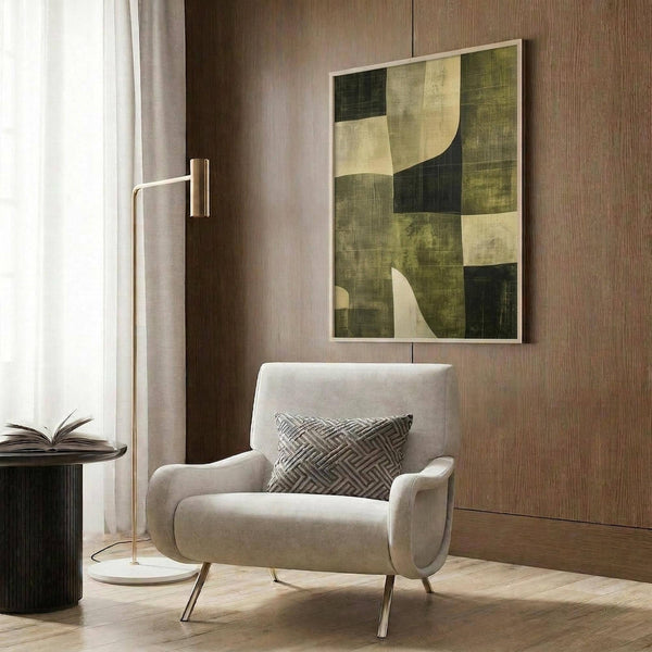 May include: Abstract geometric poster with green, cream, and dark color blocks and curved shapes, displayed in light wood frame on wood paneled wall above beige armchair with patterned pillow, brass floor lamp, and dark round side table with open book in modern neutral living room setting.