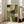 May include: Abstract geometric artwork with earth-toned blocks in various shades of green, olive, cream, and beige with curved white shapes overlaying the composition. The piece has a textured, weathered appearance and is displayed in a light wood frame, leaning against a white wall next to a beige upholstered chair in a bright, naturally-lit room with white walls and wood floors.