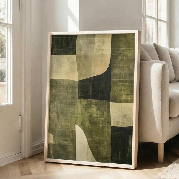 May include: Abstract geometric artwork with earth-toned blocks in various shades of green, olive, cream, and beige with curved white shapes overlaying the composition. The piece has a textured, weathered appearance and is displayed in a light wood frame, leaning against a white wall next to a beige upholstered chair in a bright, naturally-lit room with white walls and wood floors.