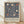 May include: Framed poster with intricate blue floral pattern featuring cream and coral tulip blooms amid swirling botanical vines. White text below reads "Centenary Exhibition William Morris 1934 Textile Industry London Jan 6 to May 6 Free Admission." Natural wood frame sits on light wooden floor against white textured wall with soft shadows.