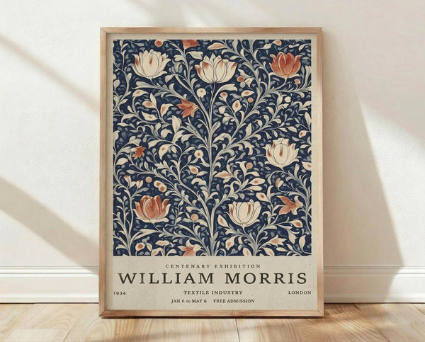 May include: Framed poster with intricate blue floral pattern featuring cream and coral tulip blooms amid swirling botanical vines. White text below reads "Centenary Exhibition William Morris 1934 Textile Industry London Jan 6 to May 6 Free Admission." Natural wood frame sits on light wooden floor against white textured wall with soft shadows.