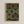 May include: Framed geometric art print with concentric circles in earth tones - browns, greens, and yellows arranged in a grid pattern against dark background. The circles have radiating lines creating a retro, mid-century modern aesthetic. Print shows "bauhaus 1919" text at bottom. Light wood frame hangs on white textured wall.