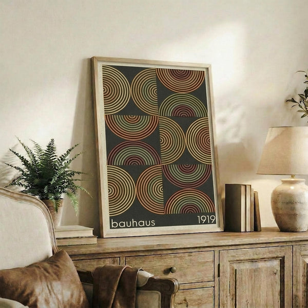 May include: A framed geometric art print featuring overlapping circles in earth tones of rust, sage green, and cream against a dark background, displayed on a rustic wooden sideboard alongside books, pottery, and a fern plant in a cozy living room setting with neutral walls and leather furniture.