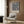 May include: Framed William Morris botanical poster with blue background and cream, orange tulip florals hanging on wood-paneled wall above beige upholstered armchair with geometric pillow. Modern brass floor lamp and dark round side table with open book complete the neutral-toned reading nook with curtained window.