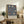 May include: Framed William Morris exhibition poster with intricate navy blue floral pattern featuring cream and coral tulips and botanical motifs, displayed on rustic wooden sideboard alongside fern plant, ceramic vases, and books in warm-lit living room with beige armchair and neutral walls
